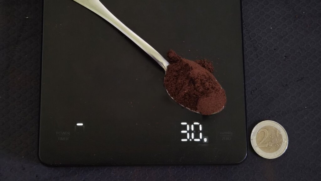 Czubata łyżeczka kawy mielonej na wadze kuchennej wskazującej 3,0 g, obok moneta 2 euro dla skali