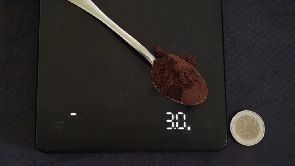 Czubata łyżeczka kawy mielonej na wadze kuchennej wskazującej 3,0 g, obok moneta 2 euro dla skali