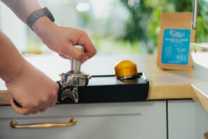 Ubijanie kawy tamperem 58 mm w kolbie, obok dystrybutor kawy i paczka kawy ziarnistej