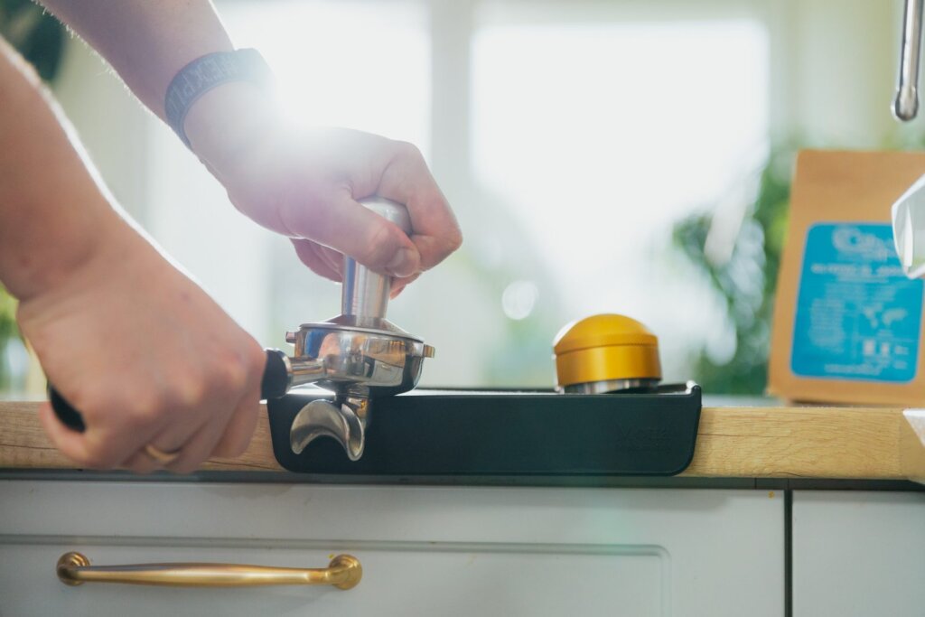 Barista ubija kawę tamperem w kolbie ekspresu, obok dystrybutor kawy na macie